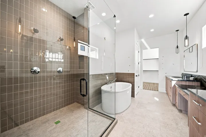 Walk-in shower with tiled walls and glass enclosure, separate oval soaking tub, dual vanity with undermount sinks and dark countertops, wood-finish cabinetry, and recessed lighting