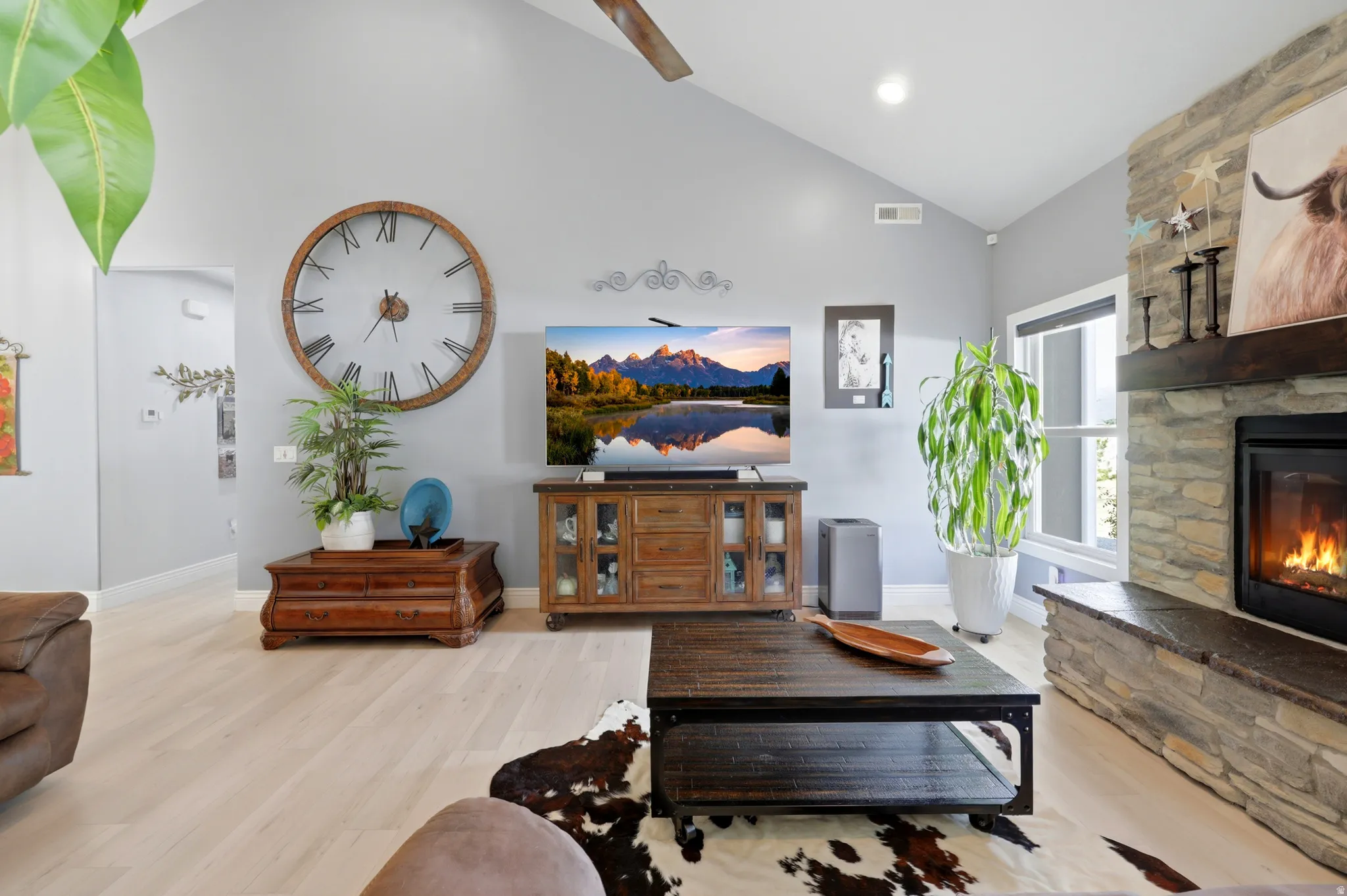 Vaulted ceiling living area featuring light wood-finish flooring, a stone-front fireplace with a wood mantel, a large window, and light grey wall paint