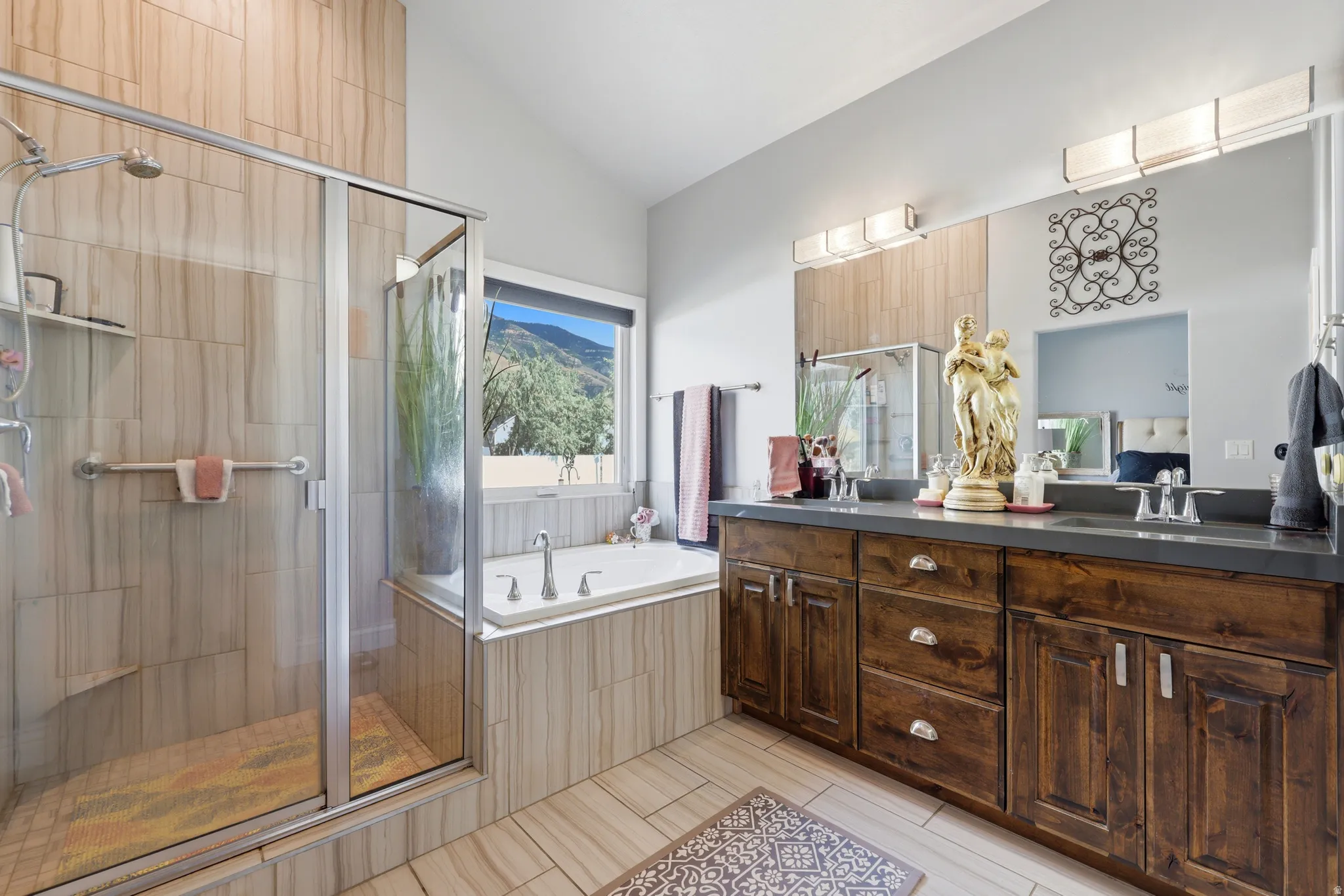 Spacious bathroom featuring a glass-enclosed shower with tile surround, a built-in soaking tub with mountain scenery, a dual-sink vanity with dark wood cabinetry, and extensive tile flooring