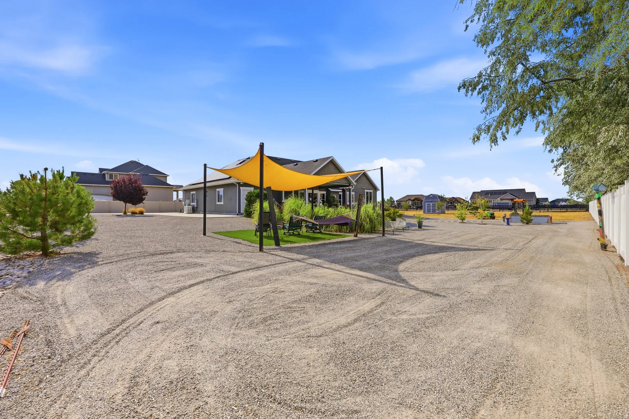 Expansive gravel lot featuring a sunshade structure, a manicured lawn area, and mature trees