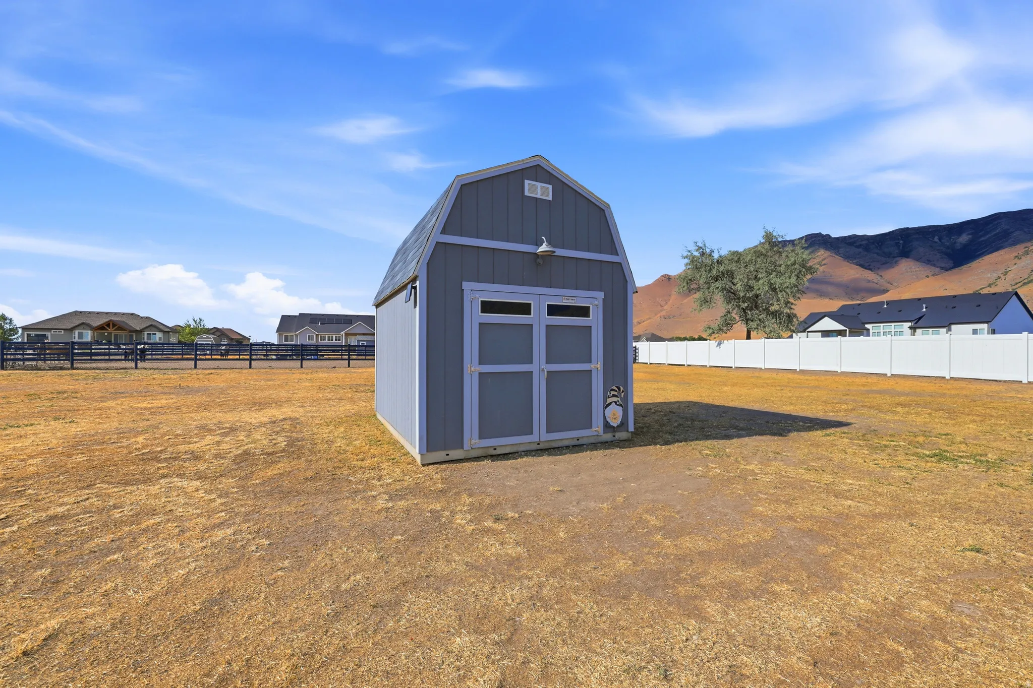 Detached storage shed with a gambrel roof and light gray siding, featuring a mounted exterior light fixture