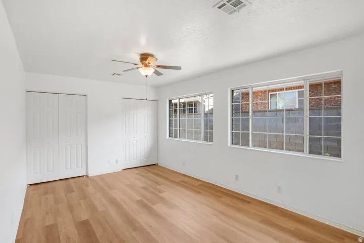 Bright interior space featuring wood-finish flooring, two large windows, a ceiling fan with integrated lighting, and two sets of bi-fold closet doors