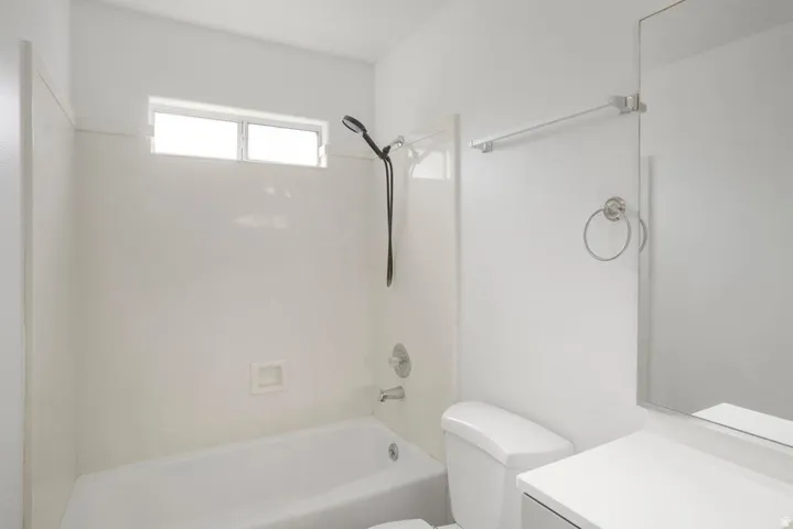 Bathroom featuring a white tub and shower combination with a window, a toilet, a vanity with a white countertop, and a wall-mounted mirror