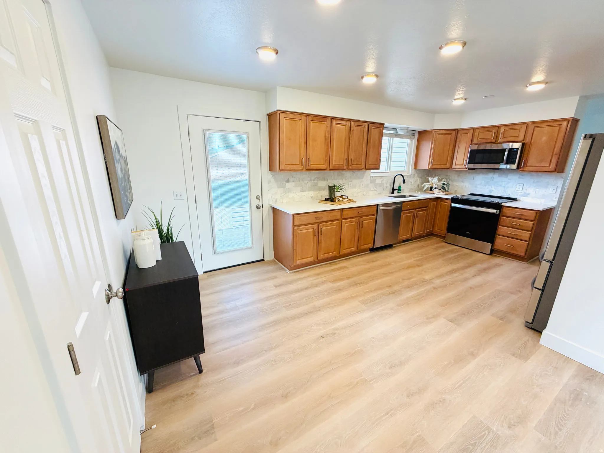 Open-concept kitchen featuring wood-finish flooring, light wood cabinetry, stone-finish countertops, stainless steel appliances, and a tile backsplash