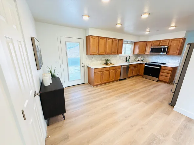 Open-concept kitchen featuring wood-finish flooring, light wood cabinetry, stone-finish countertops, stainless steel appliances, and a tile backsplash