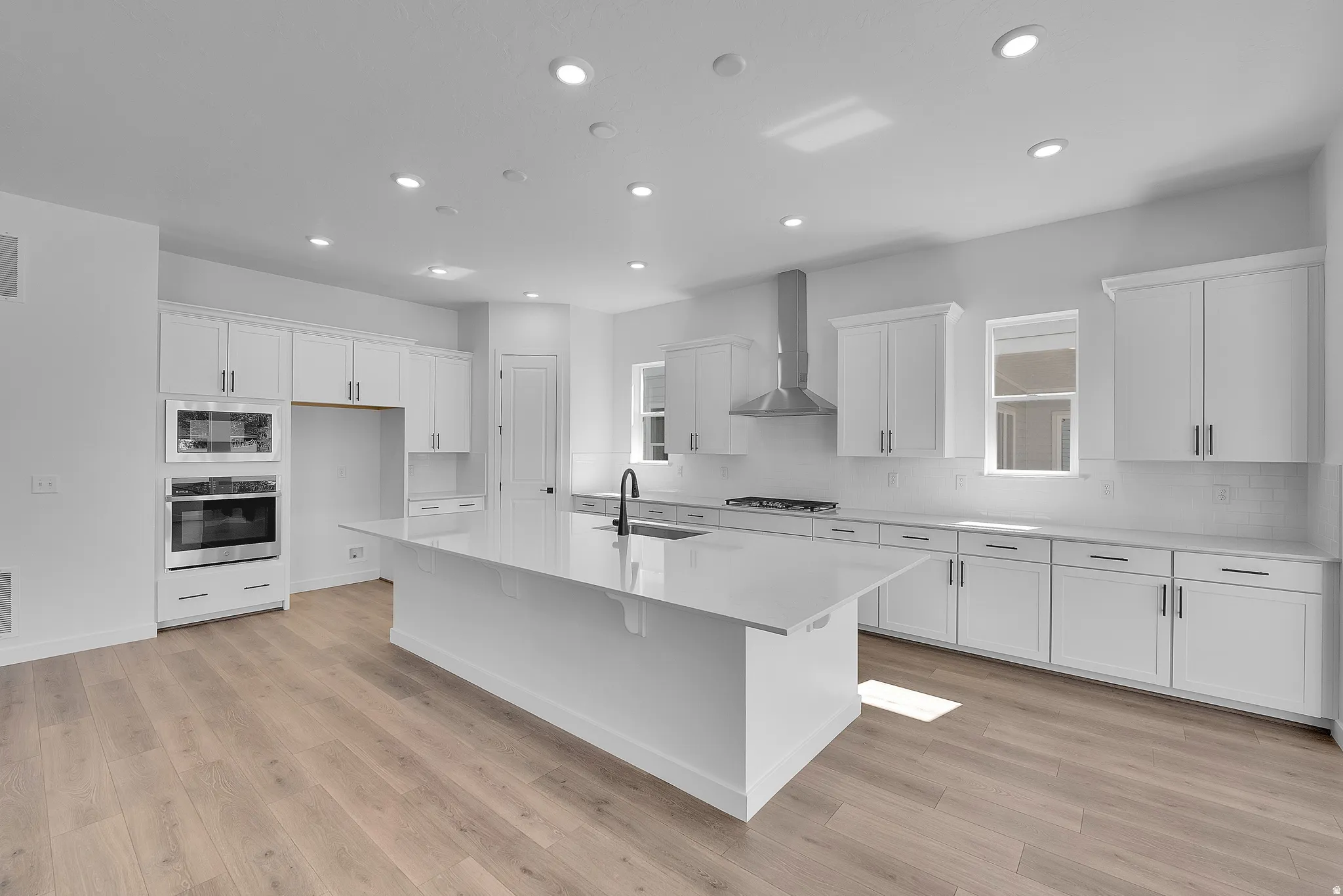 Spacious kitchen featuring an expansive central island with a sink, white cabinetry with dark hardware, built-in stainless steel appliances, wood-finish flooring, and recessed lighting