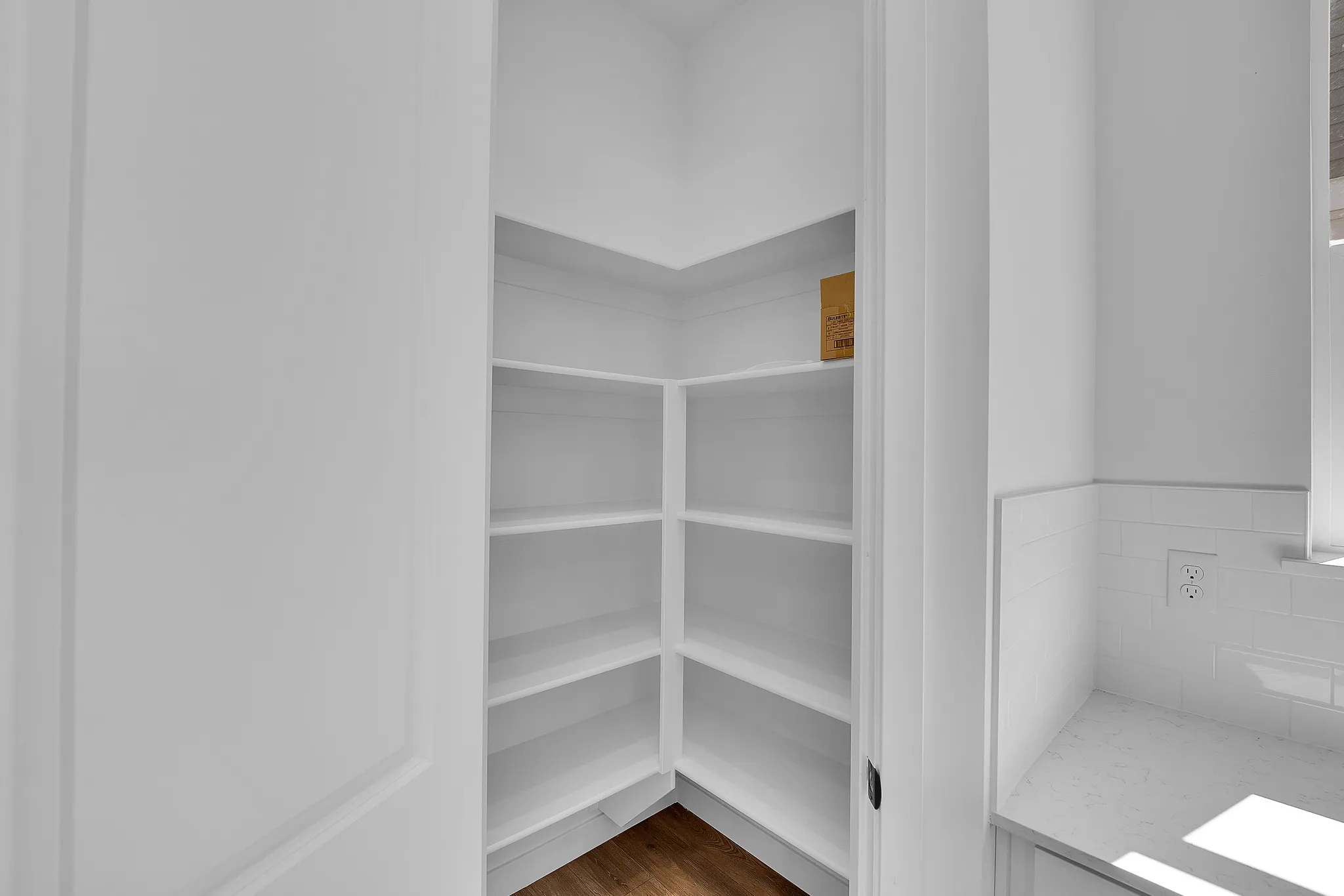 Walk-in corner pantry with multiple shelving units, wood-finish flooring, and bright white walls