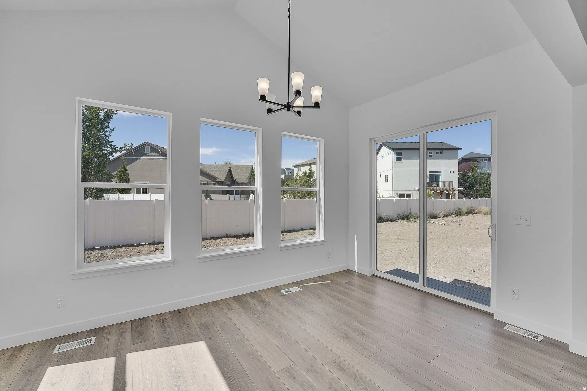 Spacious room featuring vaulted ceilings, three large windows, light wood-finish flooring, a contemporary chandelier, and direct outdoor access via a sliding glass door