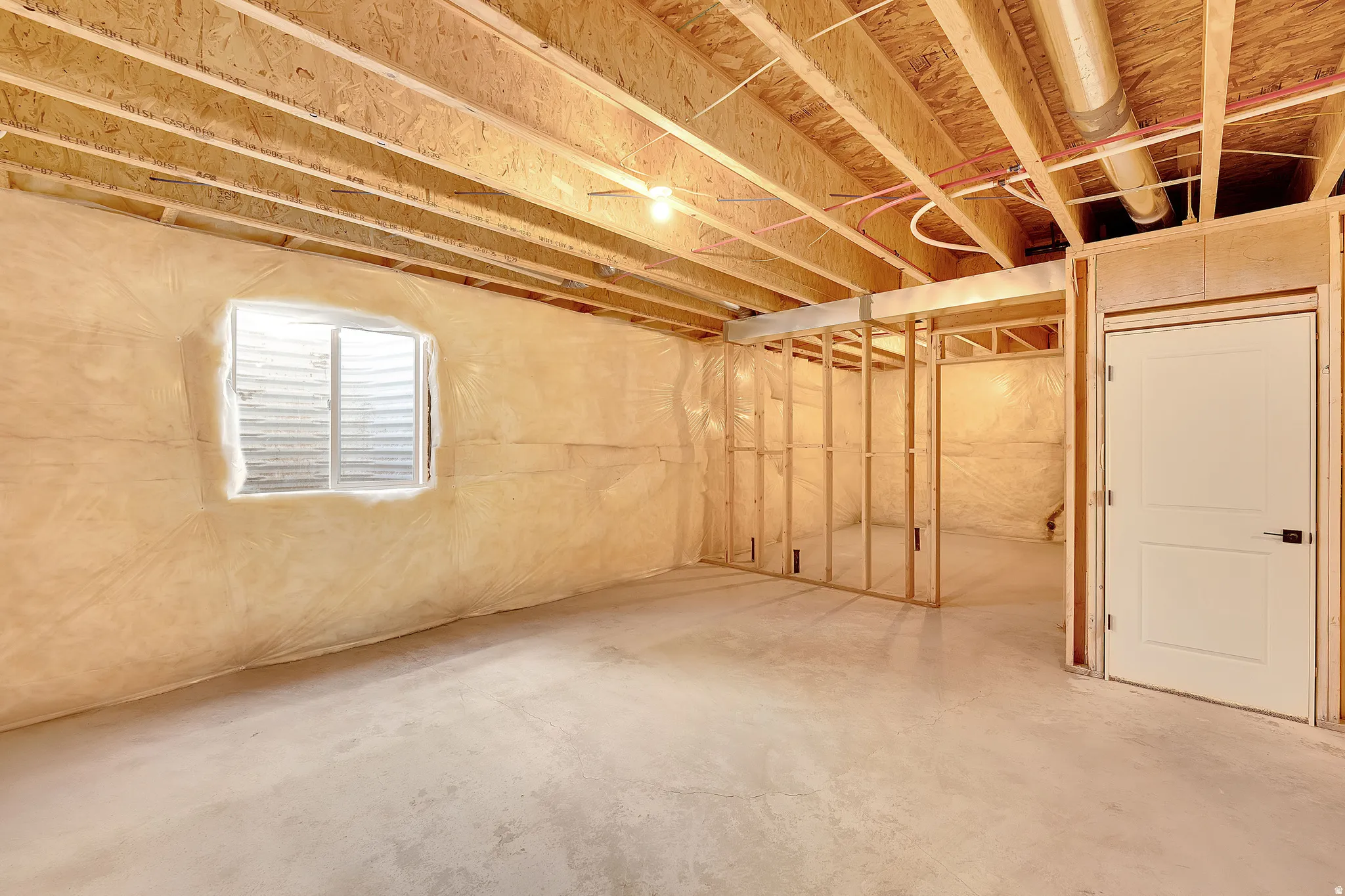 Unfinished basement featuring exposed wood joists, concrete flooring, window well, framed interior walls, and an interior door with black hardware