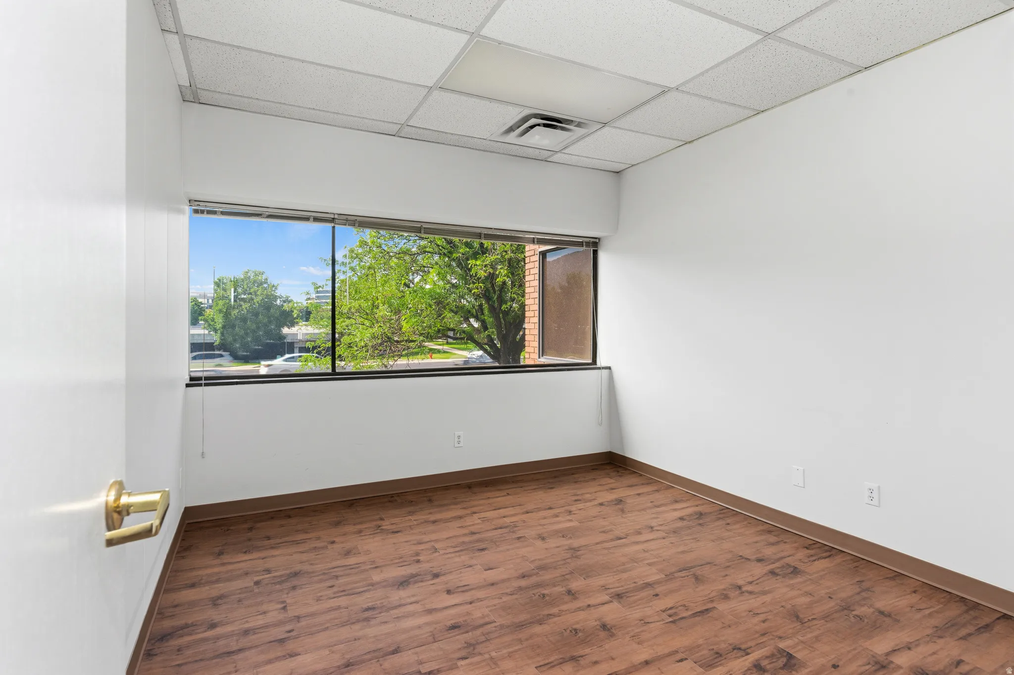 Bright interior space featuring a wide window with exterior landscaping, wood-finish flooring, white walls, drop ceiling, and brass door hardware