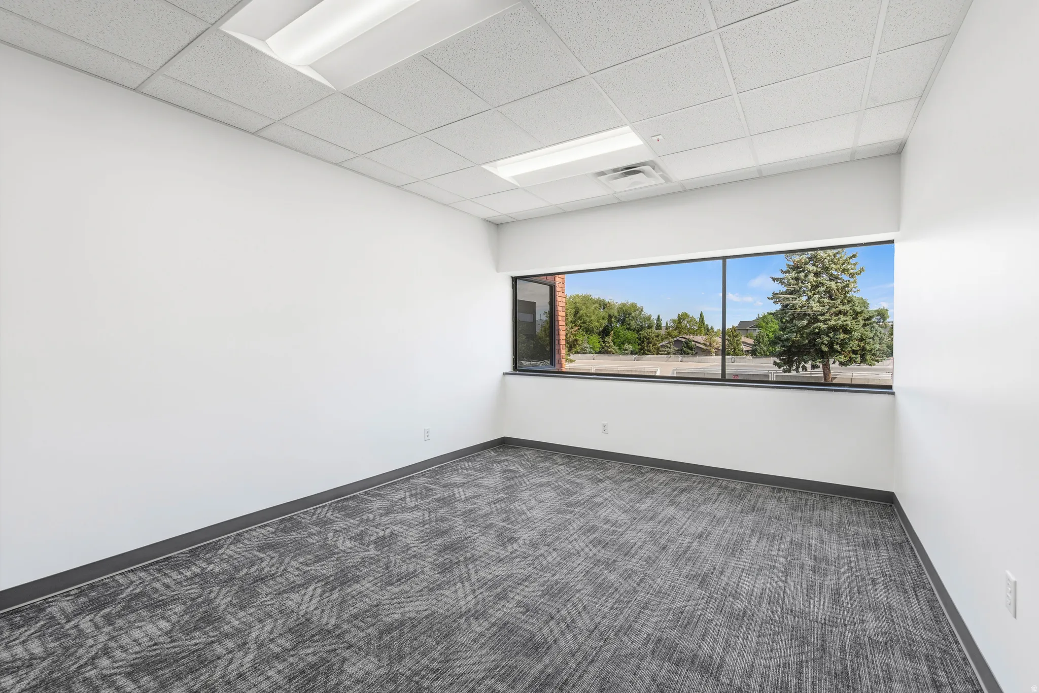 Spacious room featuring a wide window, neutral white walls, patterned carpeting, and a drop ceiling with integrated lighting