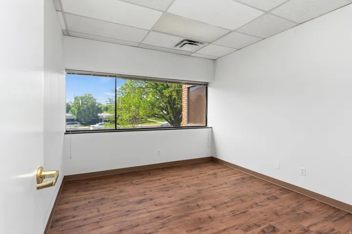Bright interior space featuring a wide window with exterior landscaping, wood-finish flooring, white walls, drop ceiling, and brass door hardware