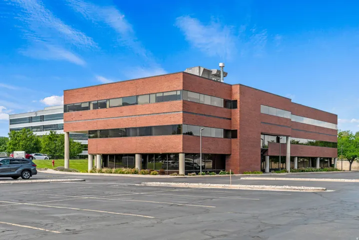Brick exterior commercial building featuring multiple stories of dark-tinted windows, a prominent entrance with supporting columns, and an expansive asphalt parking area