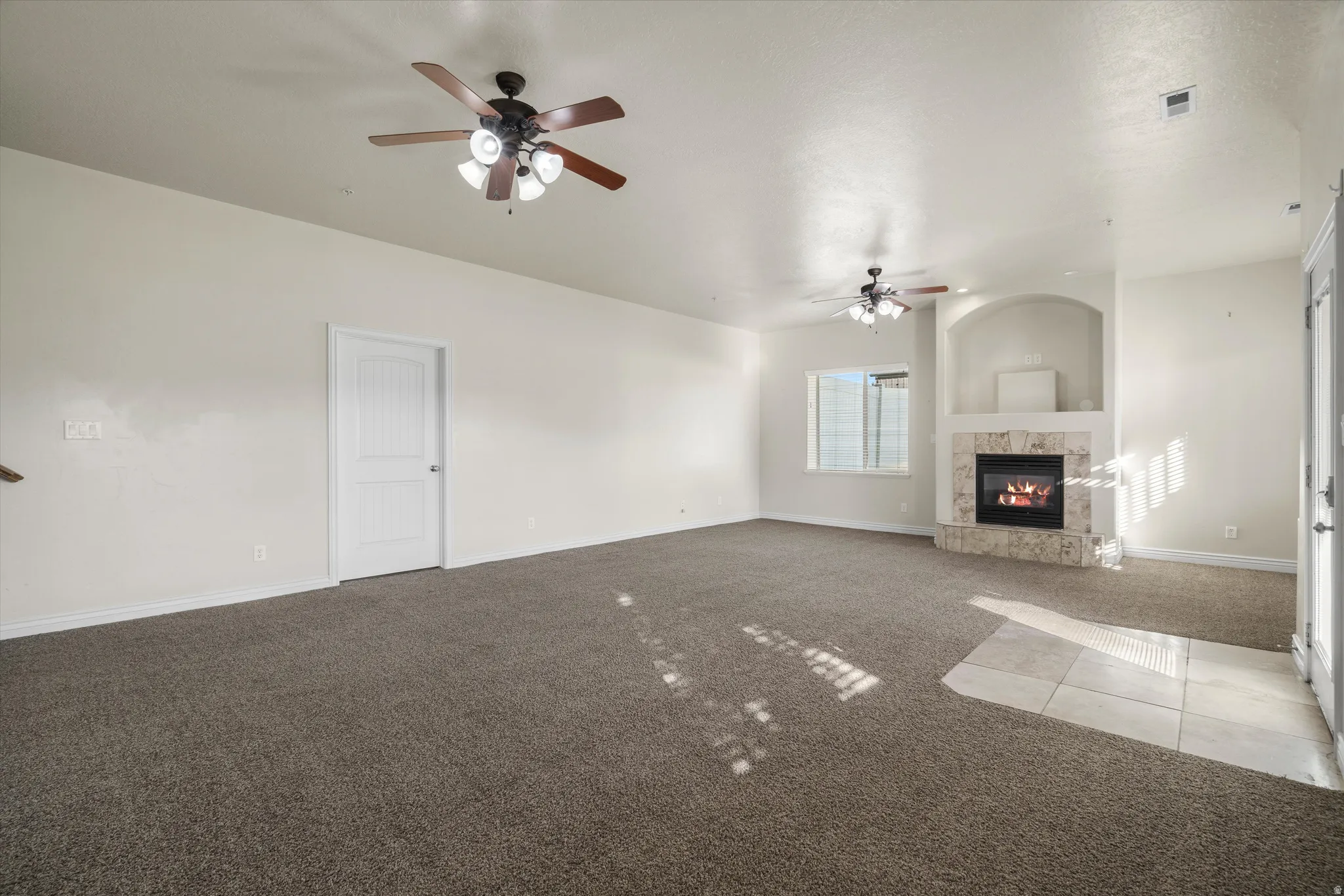 Spacious living area featuring neutral-toned carpeting, a built-in fireplace with stone surround, two ceiling fans with integrated lighting, and multiple windows