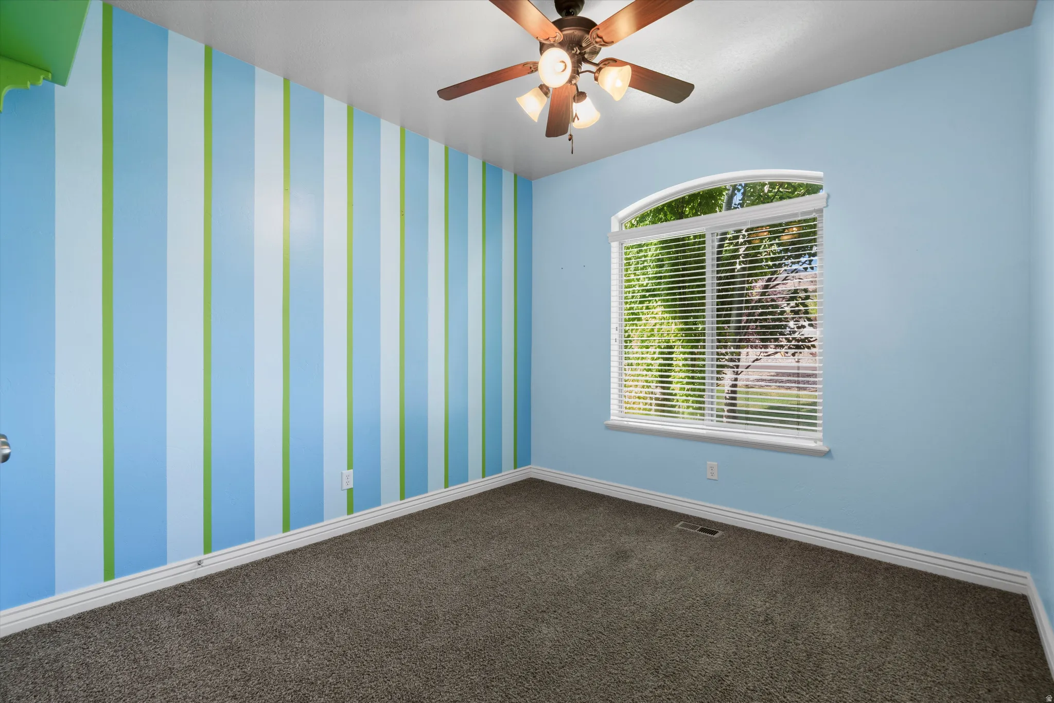 Carpeted room featuring a large arched window with horizontal blinds, a ceiling fan with integrated lighting, and a wall with blue and green vertical stripes