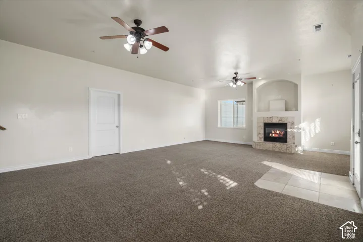 Spacious living area featuring neutral-toned carpeting, a built-in fireplace with stone surround, two ceiling fans with integrated lighting, and multiple windows
