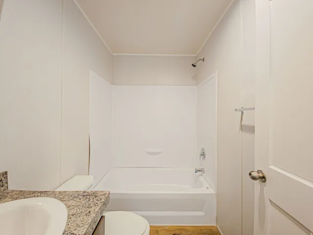 White paneled walls, white bathtub with integrated shelving, chrome shower fixture, chrome towel bar, and a wood-finish floor