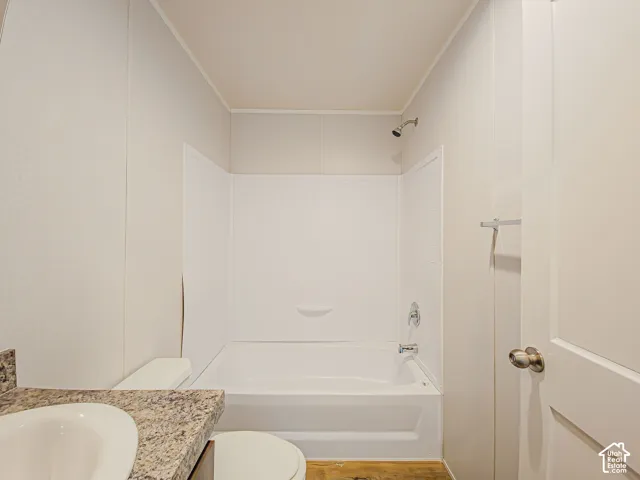 White paneled walls, white bathtub with integrated shelving, chrome shower fixture, chrome towel bar, and a wood-finish floor