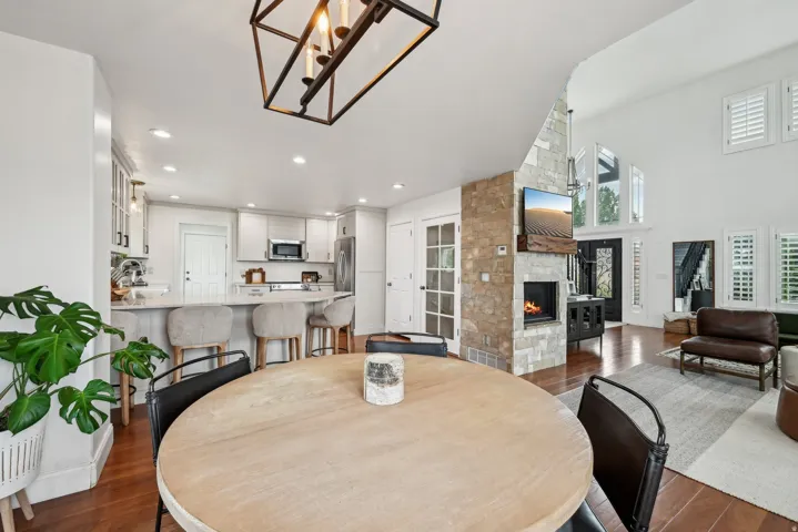 Open-concept living area featuring a stone-clad fireplace, high vaulted ceilings, wood-finish flooring, a kitchen with white cabinetry and stainless steel appliances, and a breakfast bar with seating