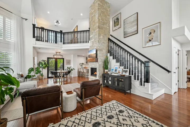Spacious living area featuring high ceilings, wood-finish flooring, a stone-clad fireplace, and a striking staircase with black balusters