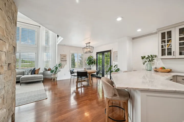 Open-concept living area featuring rich wood-finish flooring, a stone accent wall, and abundant natural light from multiple windows with plantation shutters