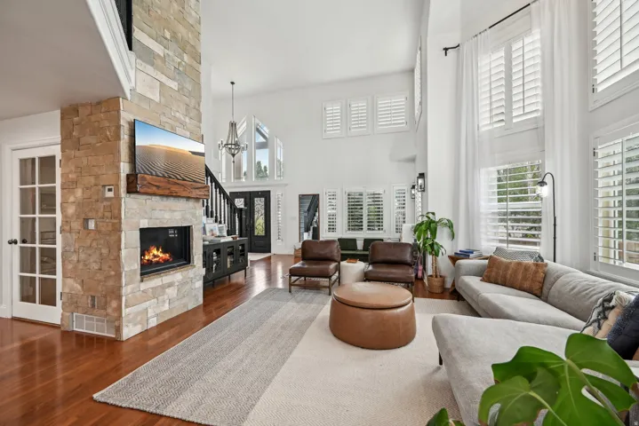 Two-story living area featuring a stone fireplace, wood-finish flooring, high ceilings, large windows with white shutters, and a chandelier