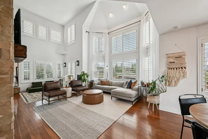 Open-concept living space featuring high vaulted ceilings, extensive window banks with plantation shutters, wood-finish flooring, a stone accent wall, and recessed lighting