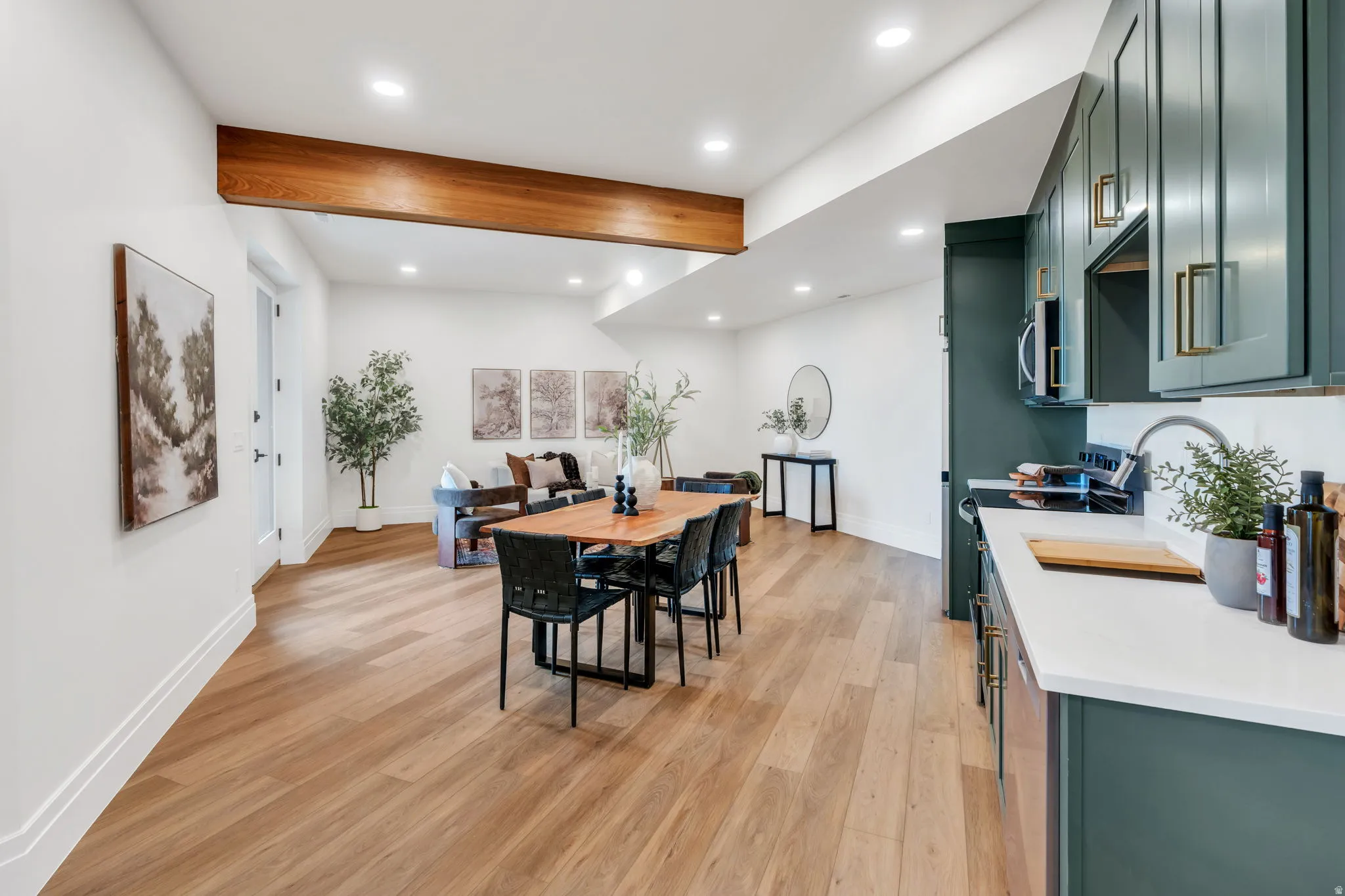 Open-concept living area featuring wood-finish flooring, a prominent exposed wood beam, recessed lighting, and a kitchen with deep green cabinetry and a white countertop