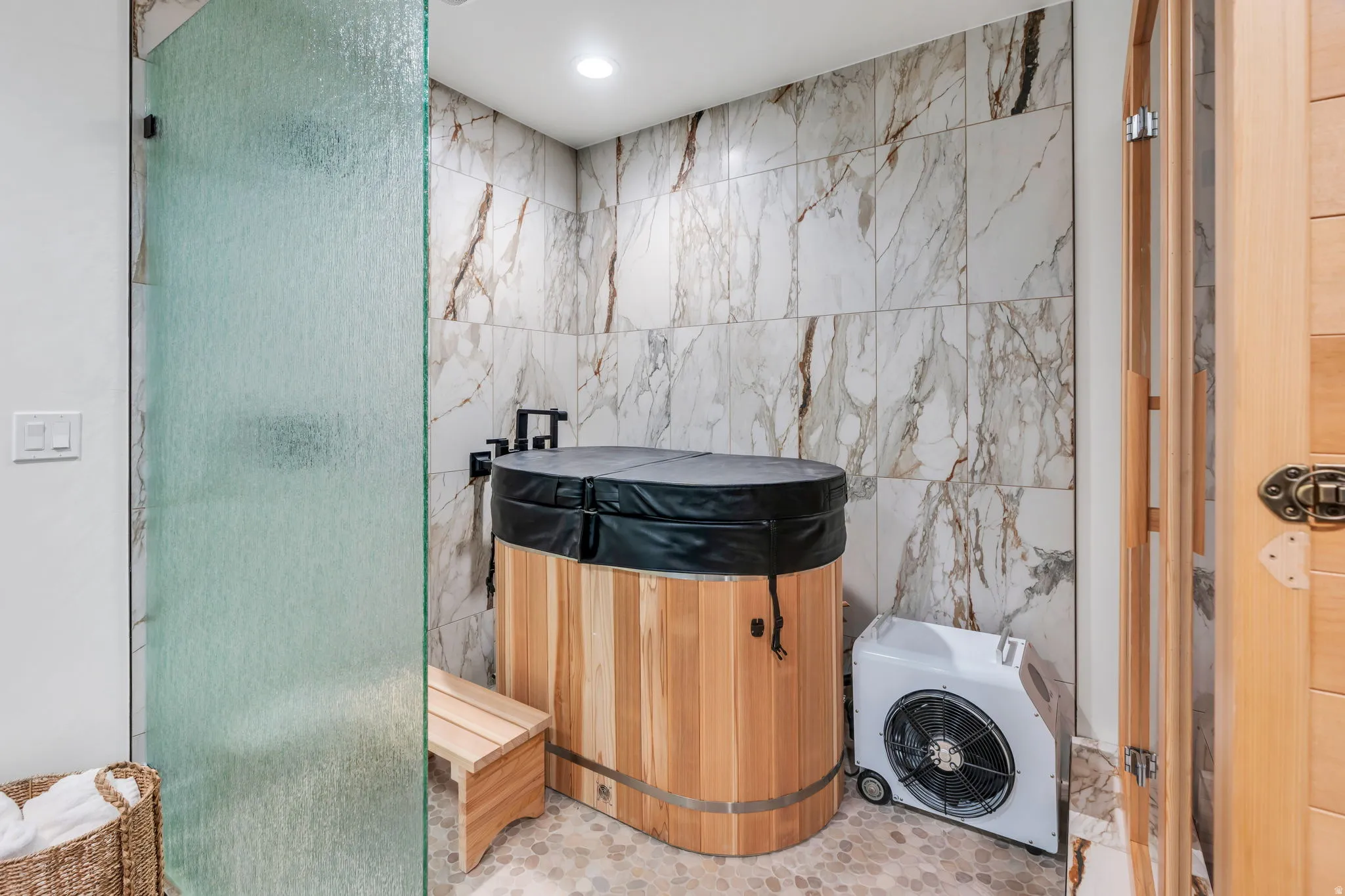 Contemporary spa-inspired bathroom featuring a wooden soaking tub with black cover, large format marble-look wall tiles, and pebble-finish floor tiles