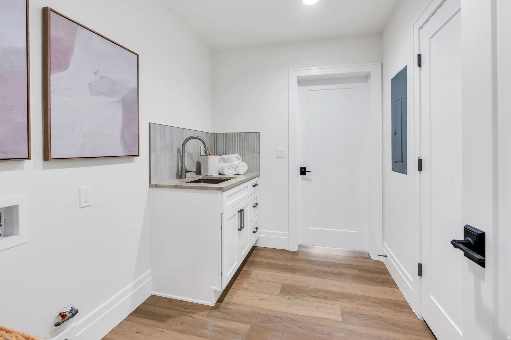 Laundry area featuring wood-finish flooring, white cabinetry with black hardware, a stainless steel sink with a gooseneck faucet, a light-toned countertop, and a tiled backsplash