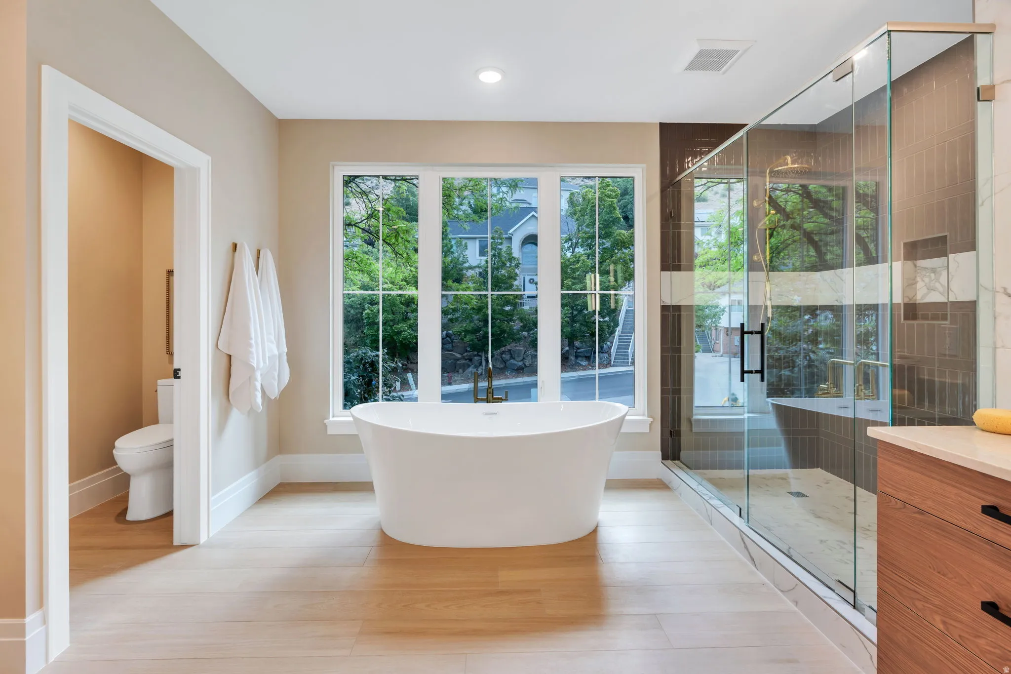 Contemporary bathroom featuring a freestanding soaking tub, expansive window, glass-enclosed shower, wood-finish flooring, and a separate water closet
