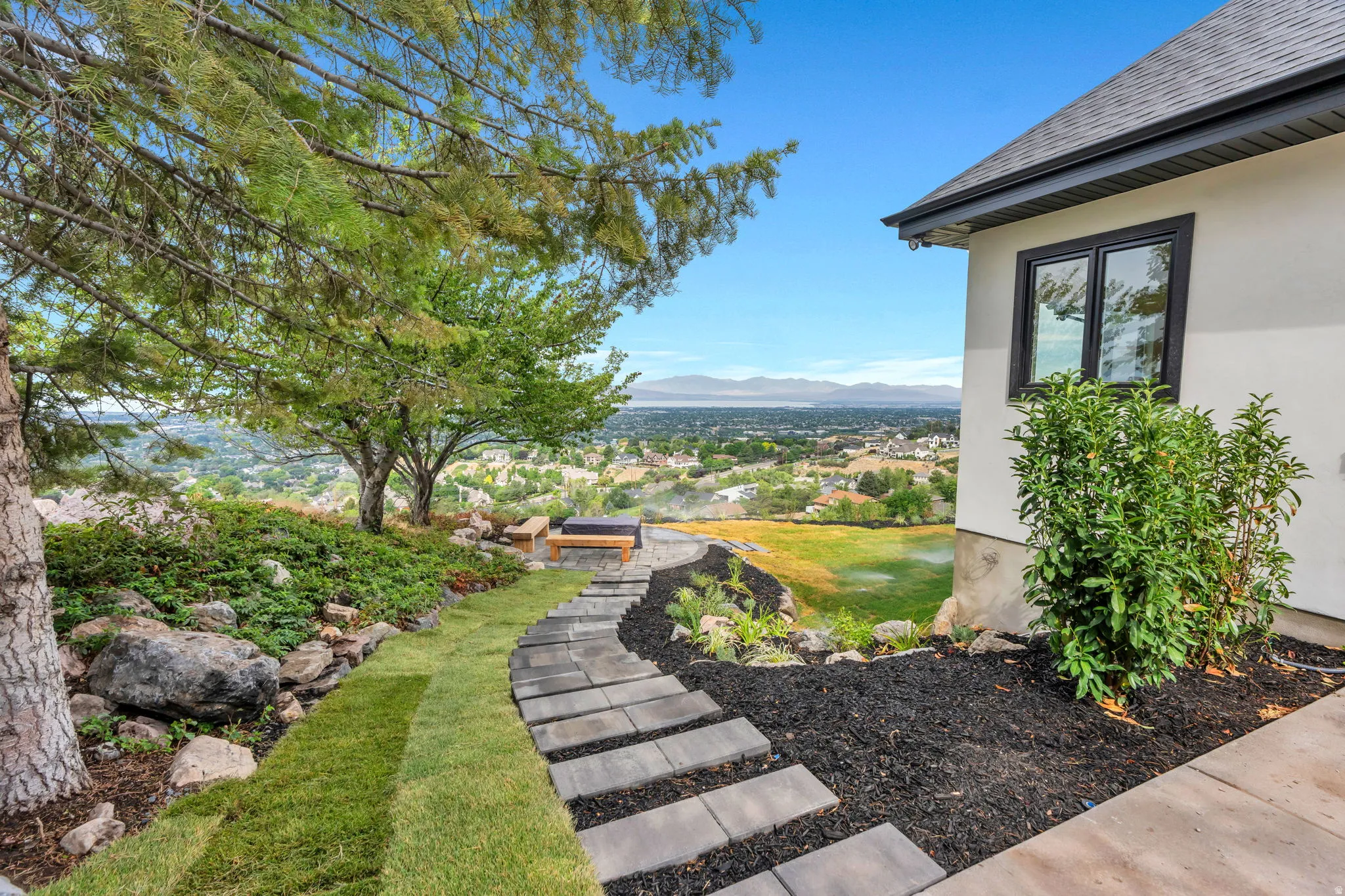 Terraced landscaping featuring a stone paver pathway, mature trees, manicured lawn, and planted garden beds