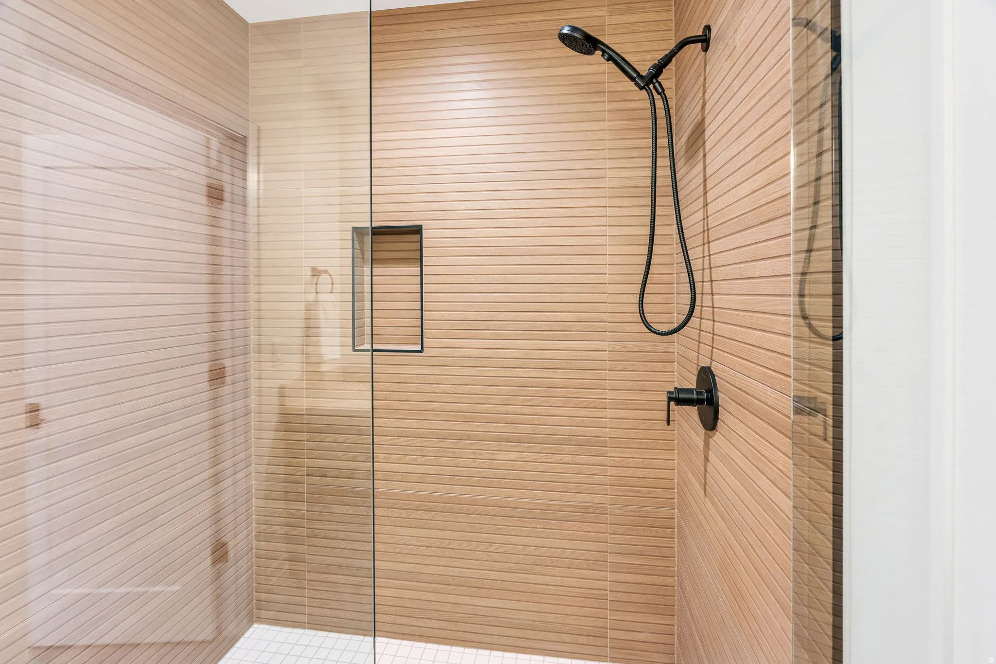 Shower enclosure with glass paneling, horizontal textured tile, a black matte handheld showerhead, a recessed shower niche, and a contrasting white tiled floor