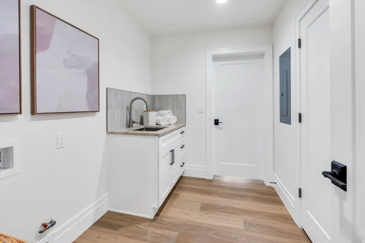 Laundry area featuring wood-finish flooring, white cabinetry with black hardware, a stainless steel sink with a gooseneck faucet, a light-toned countertop, and a tiled backsplash