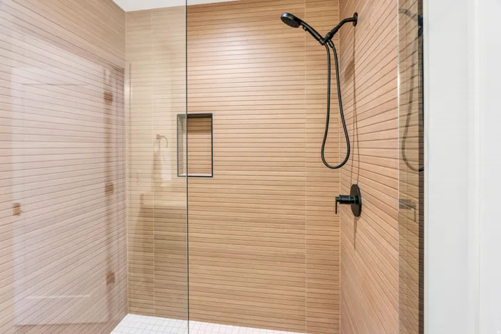 Shower enclosure with glass paneling, horizontal textured tile, a black matte handheld showerhead, a recessed shower niche, and a contrasting white tiled floor