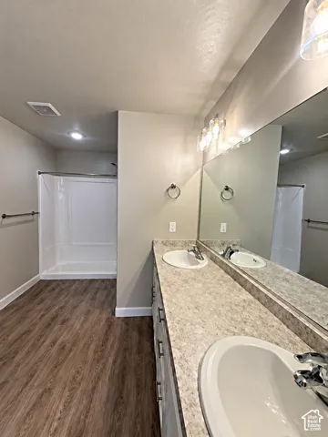 Dual vanity with integrated sinks, wood-finish flooring, full-width mirror, light-toned countertops, and a white shower/tub combination
