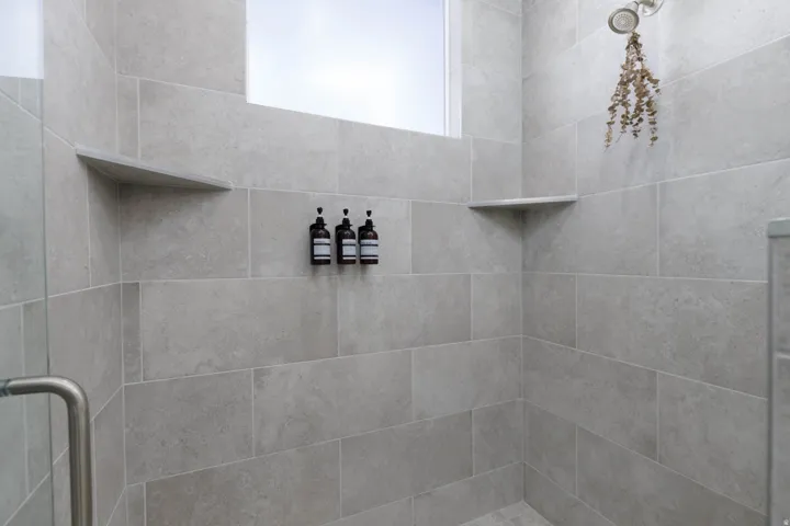 Shower stall featuring light gray rectangular tile walls, an integrated corner shelf, a built-in horizontal window, and a rain shower head