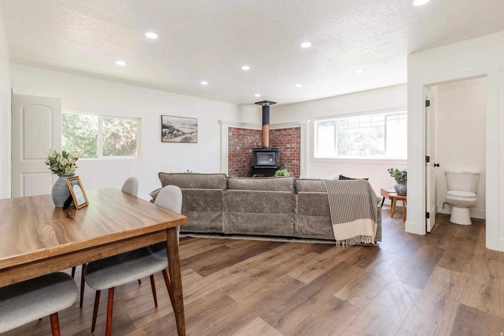 Open concept living space featuring wood-finish flooring, recessed lighting, and a brick accent wall with a wood-burning stove