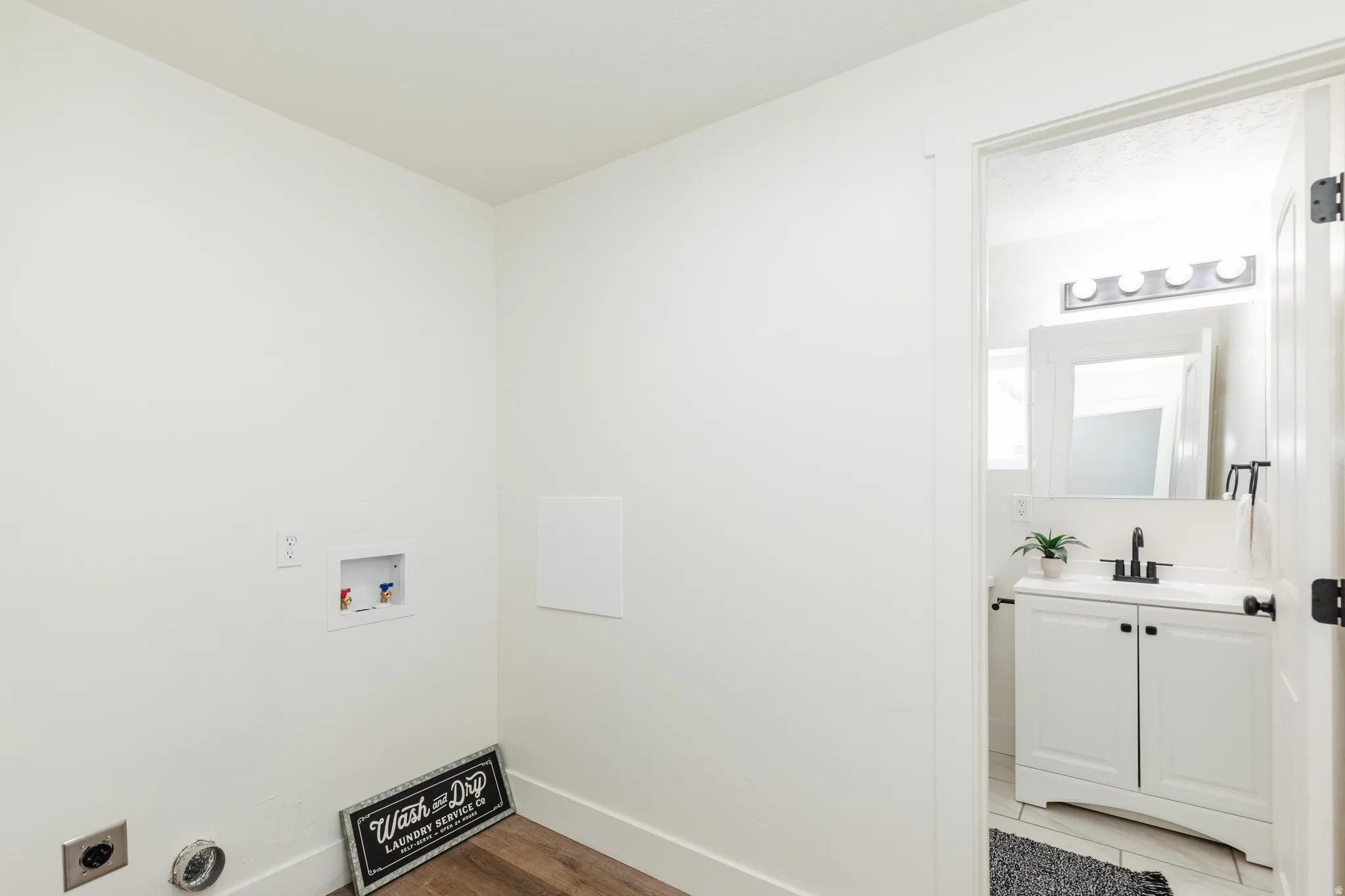 Dedicated laundry area with hookups and wood-finish flooring, opening to a bathroom featuring a white vanity with a rectangular sink, black faucet, and a wall-mounted mirror