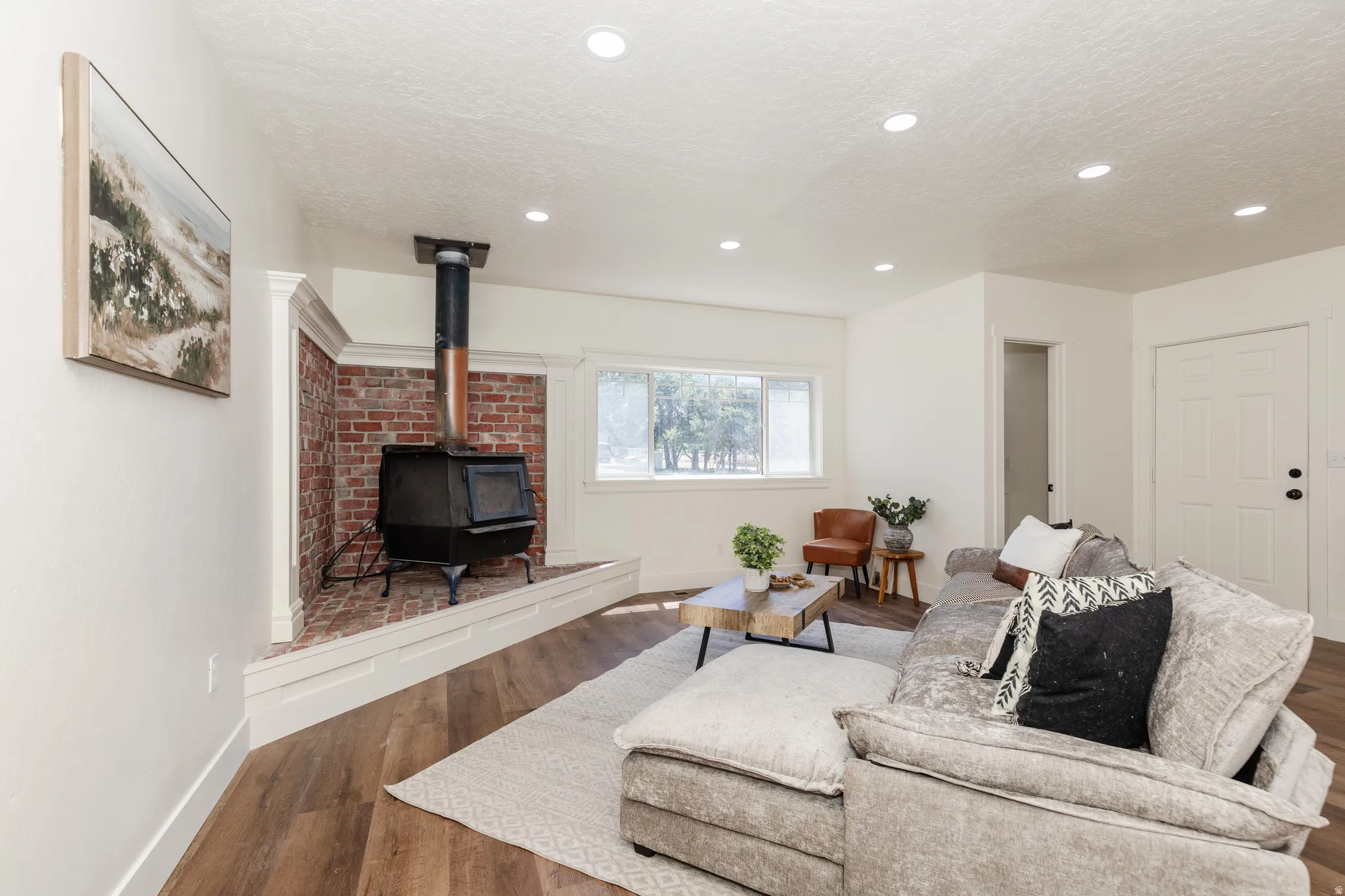 Living area featuring a brick hearth with a black wood-burning stove, recessed ceiling lighting, wood-finish flooring, and a window with exterior views