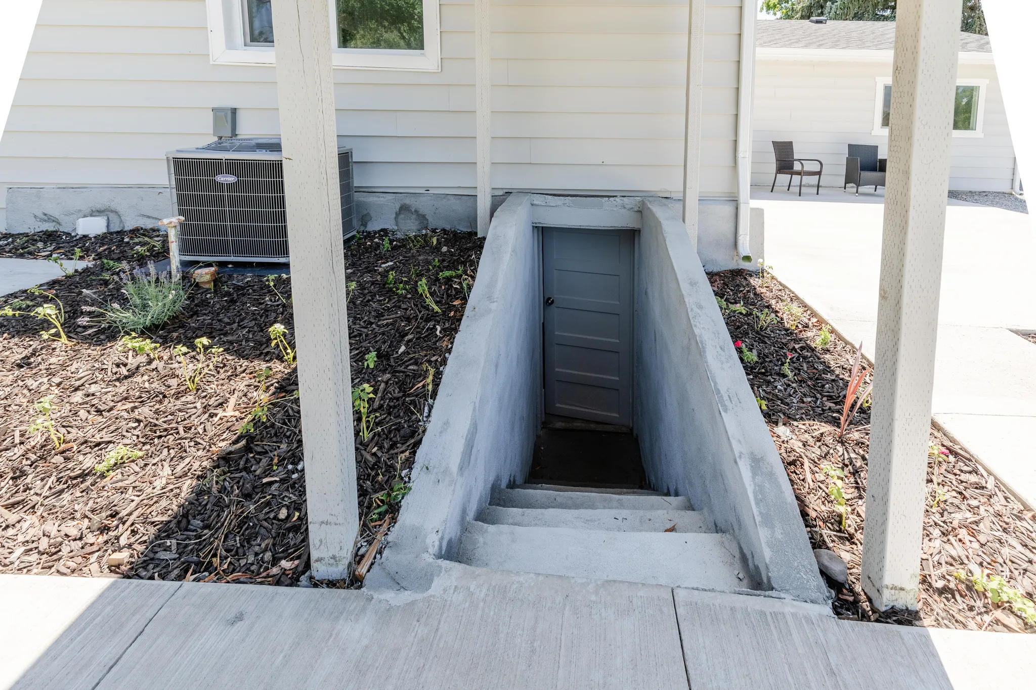 Concrete basement entryway with a solid door, flanked by mulched garden beds and a visible HVAC unit