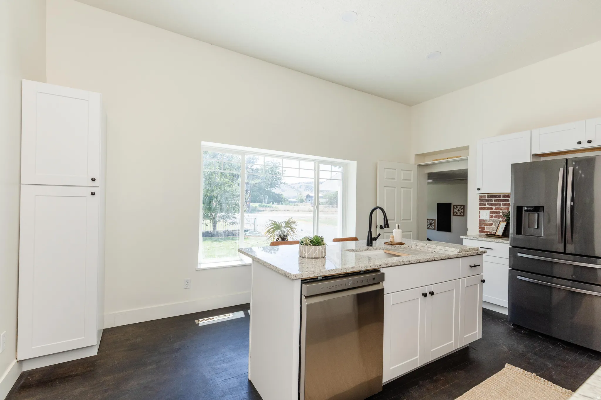 Bright kitchen featuring a central island with an undermount sink and a light-toned countertop, white cabinetry, a stainless steel dishwasher, and a dark-finish refrigerator