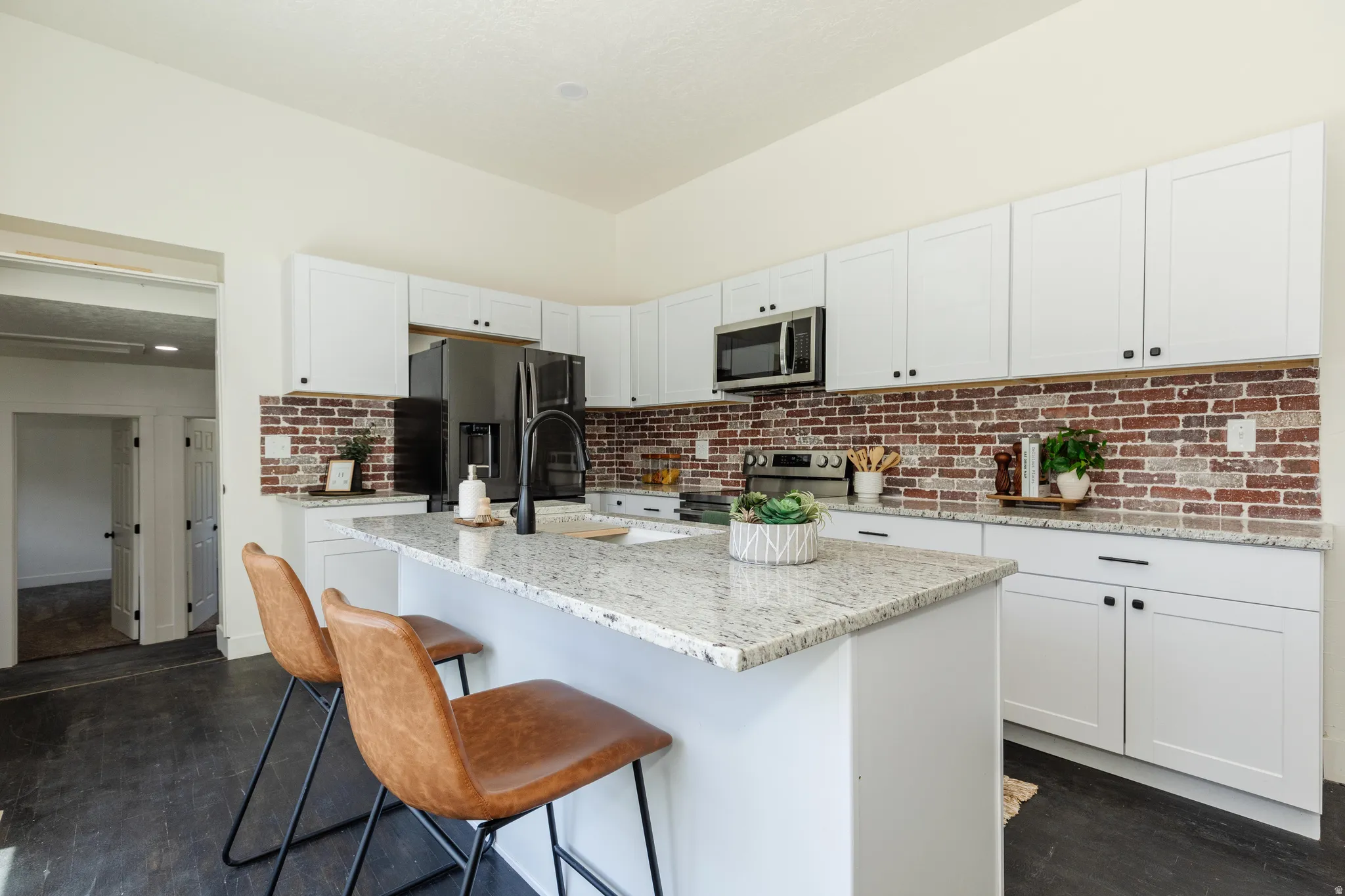 Modern kitchen featuring a central island with a light-toned granite countertop, white cabinetry with dark hardware, a brick-patterned backsplash, and dark wood-finish flooring