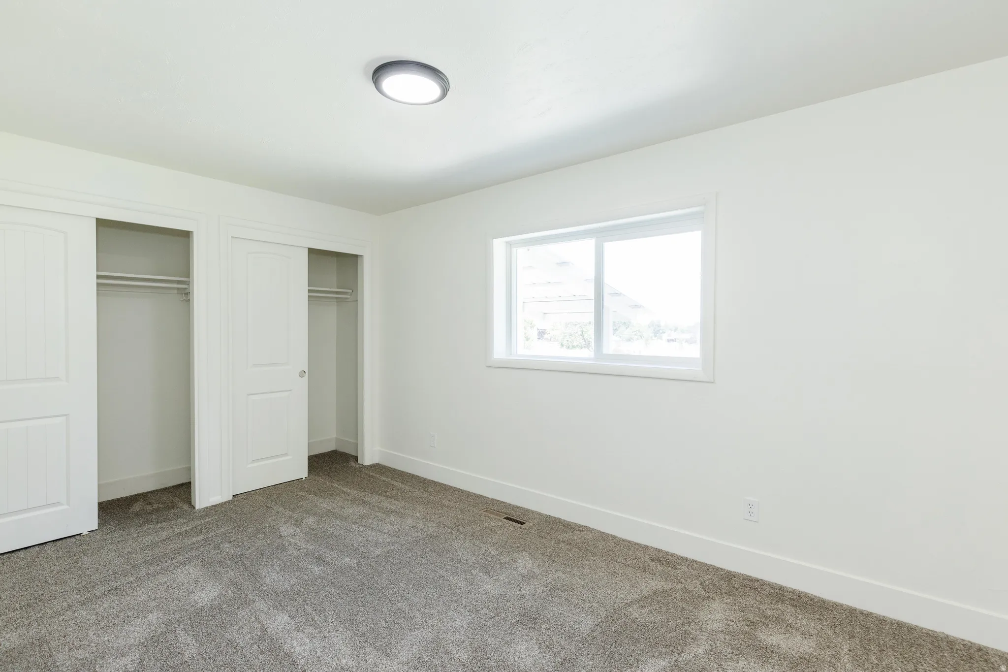Bright room featuring a window with white trim, gray carpet, white walls, and two sliding closet doors with interior shelving and hanging rods