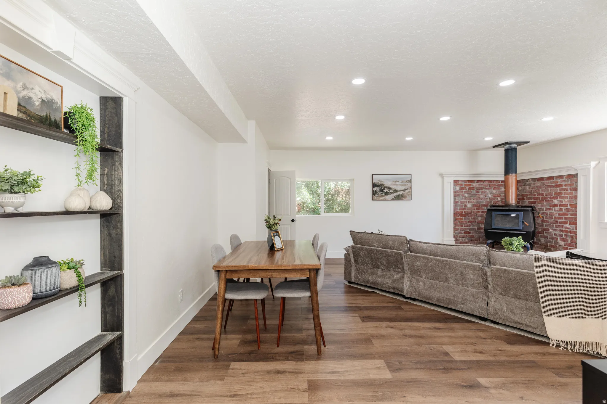 Open-concept living space featuring wood-finish flooring, recessed lighting, white walls, built-in shelving, and a brick accent wall with a wood-burning stove