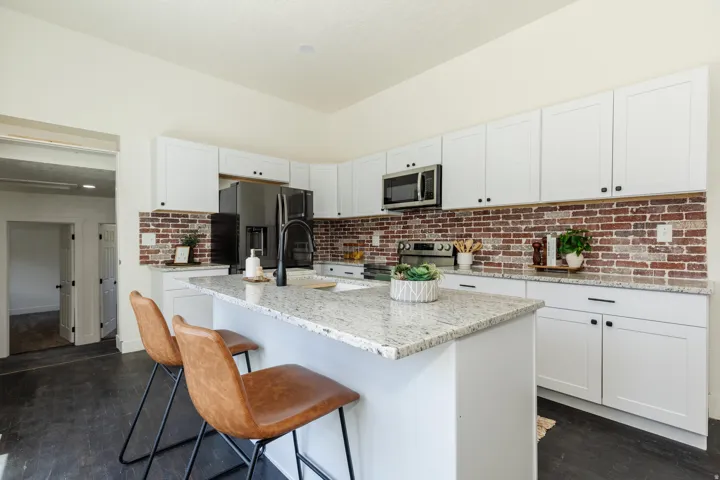 Modern kitchen featuring a central island with a light-toned granite countertop, white cabinetry with dark hardware, a brick-patterned backsplash, and dark wood-finish flooring