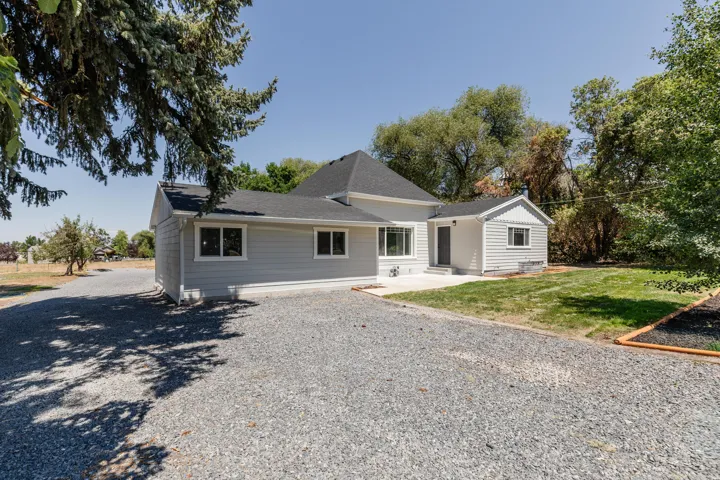 Contemporary home exterior featuring light gray siding, dark roof shingles, white trim windows, a gravel driveway, and a concrete patio
