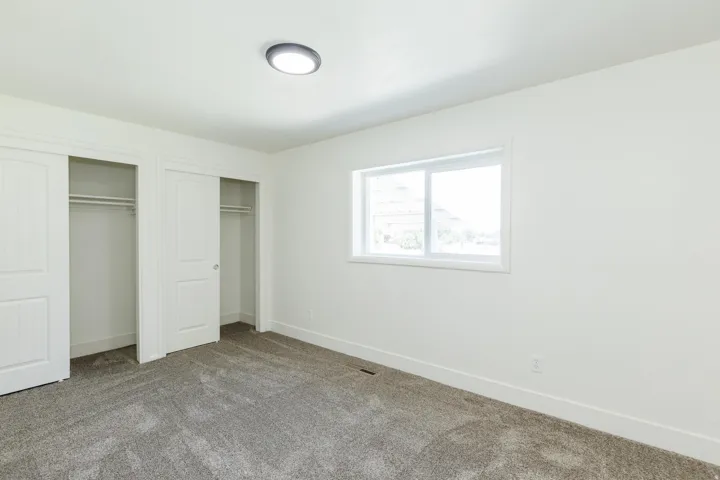 Bright room featuring a window with white trim, gray carpet, white walls, and two sliding closet doors with interior shelving and hanging rods