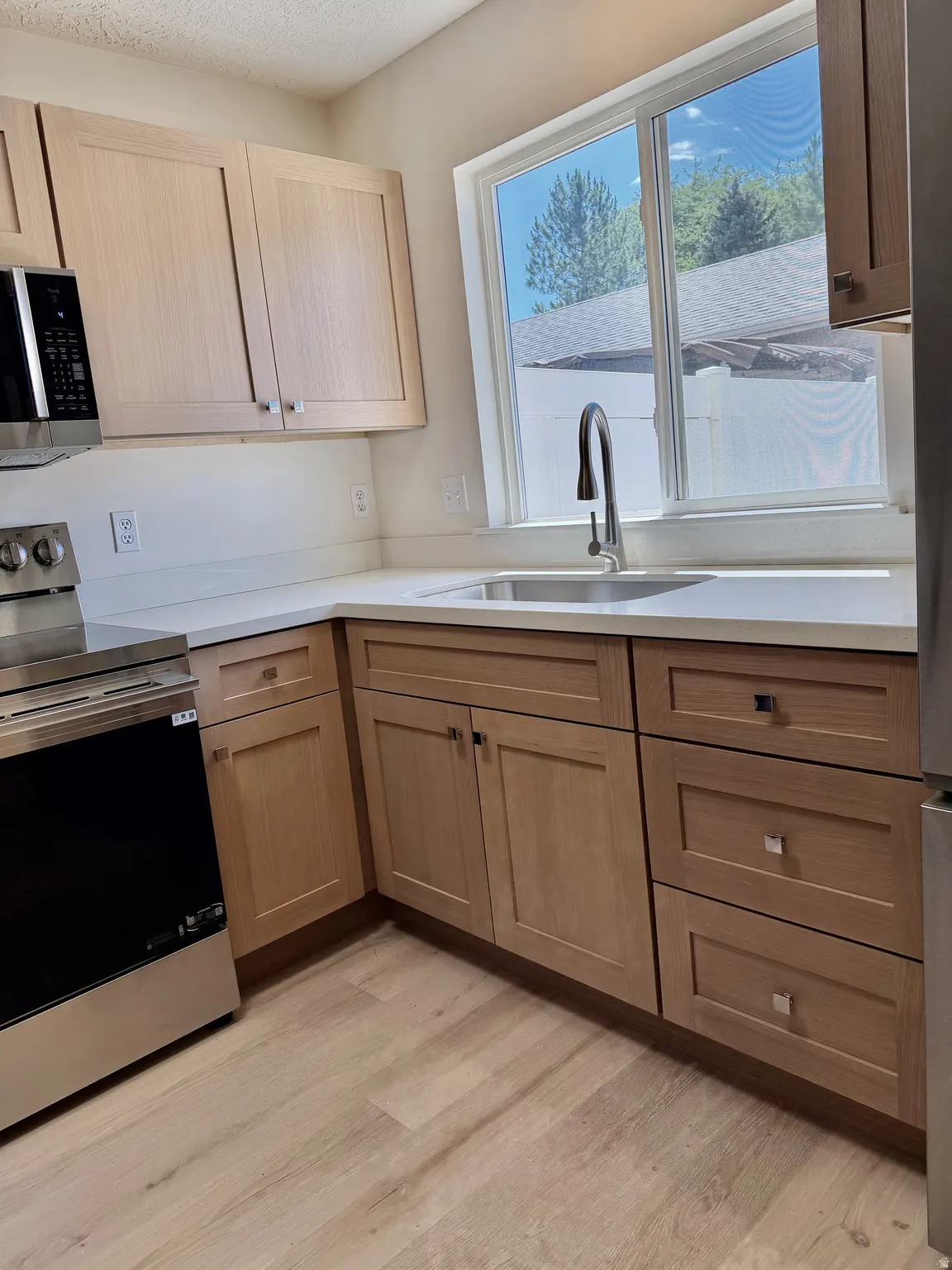 Modern kitchen featuring light wood-finish cabinetry, stainless steel appliances, a deep single-basin sink with a high-arc faucet, light-toned countertops, and wood-finish flooring