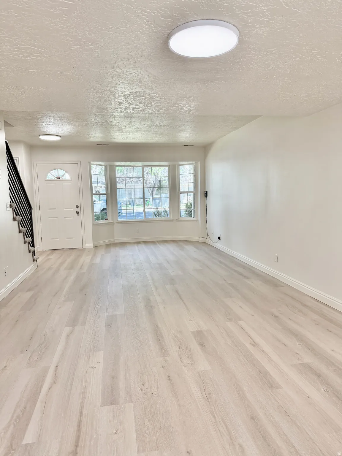 Bright living space featuring light wood-finish flooring, a bay window, and a white entry door with an arched glass insert